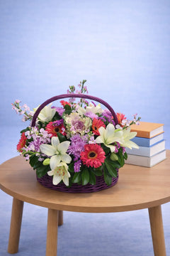 Mixed flower basket with white lilies, red gerbera daisies and purple carnations in a purple wicker basket with arched handle