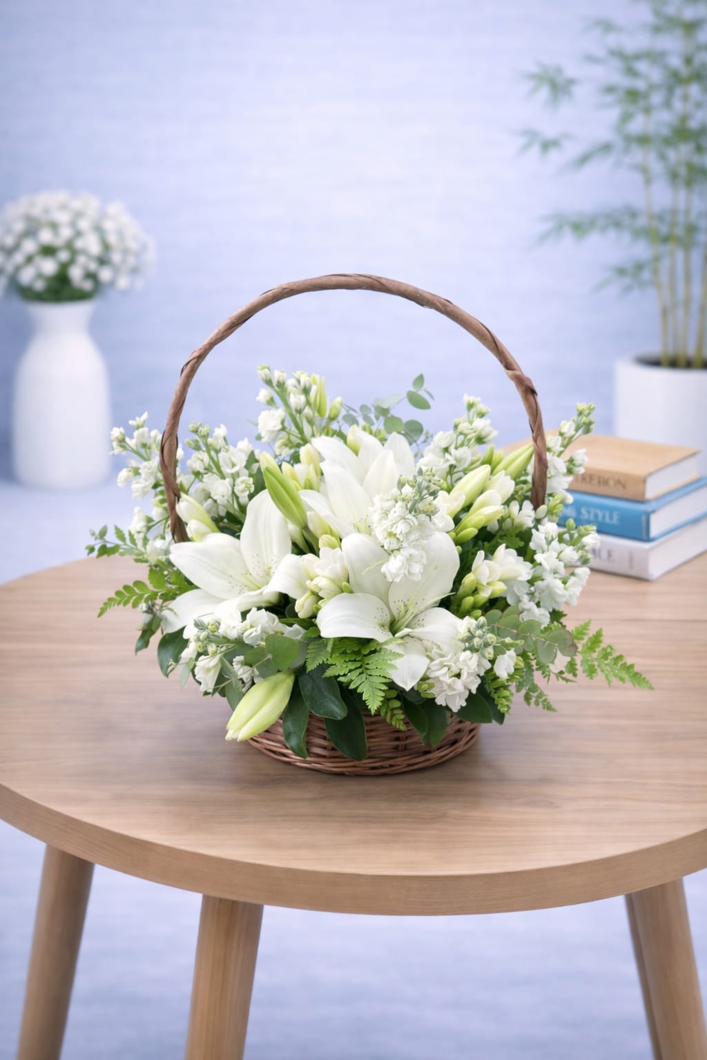 White oriental lily and stock flower arrangement in a natural wicker basket with arched handle and green fern leaves